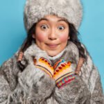 Happy woman from Alaska looks with surprised delighted face at camera wears winter hat fur coat and mittens poses against blue background. Eskimo girl dressed in outerwear for chilling frosty weather