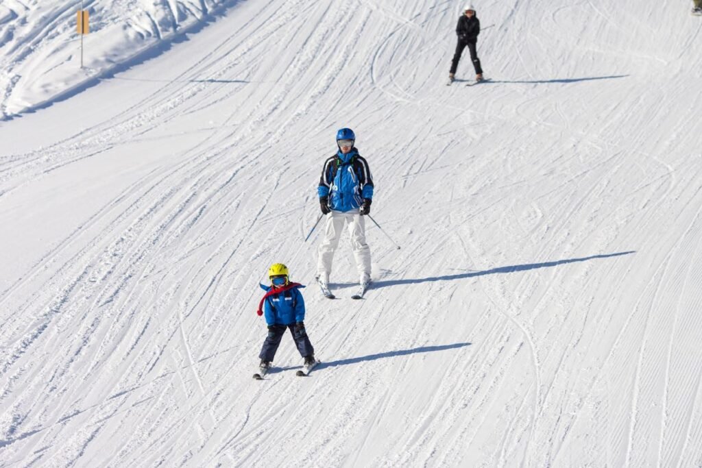 Adult skier and young child in bright ski gear learning to ski together on a groomed beginner run at a British Columbia alpine resort