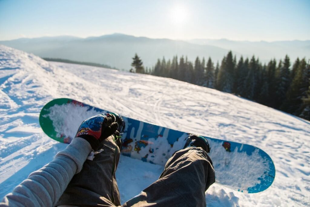 Point-of-view shot of snowboarder's legs and colorful snowboard gliding down a sunny slope with evergreen forest and mountains visible in the distance at a BC resort