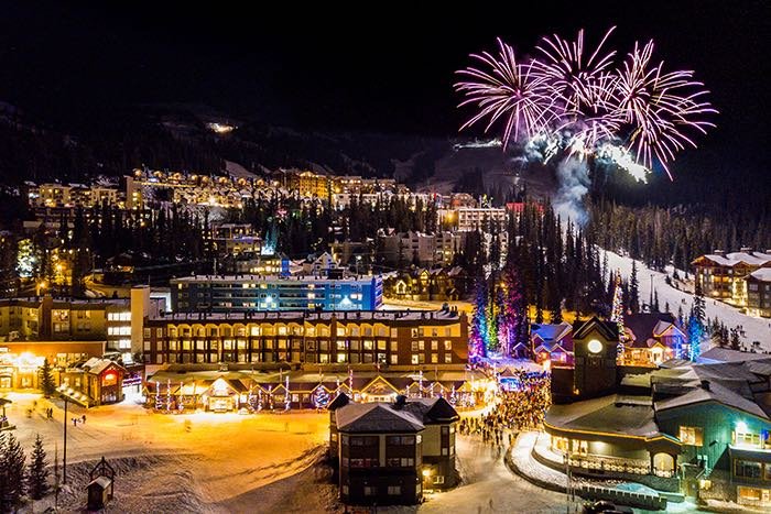 Skiing the Thompson Okanagan at Big White Ski Resort during a nighttime celebration with spectacular fireworks bursting over the illuminated village, showcasing the resort's festive alpine atmosphere and vibrant après-ski scene