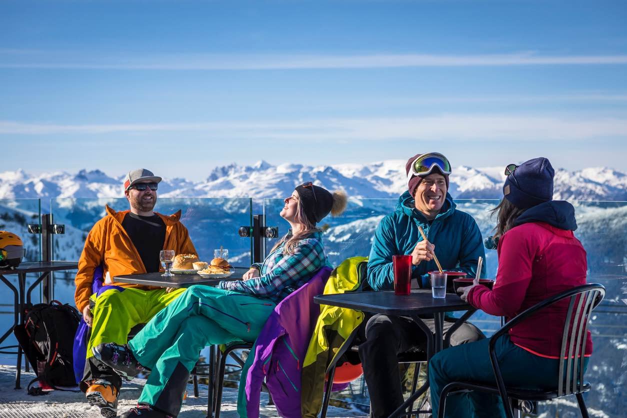 Group of skiers and snowboarders enjoying food and drinks on sunny mountain-top patio with panoramic views of snow-capped peaks at Whistler Blackcomb