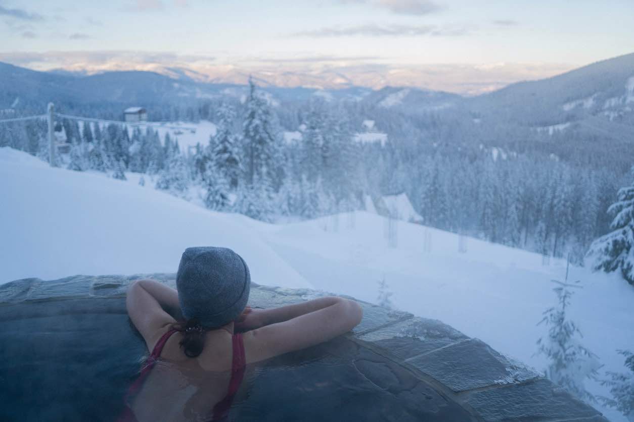 Person relaxing in outdoor infinity hot tub overlooking snowy mountain slopes and forest landscape in Whistler during winter twilight