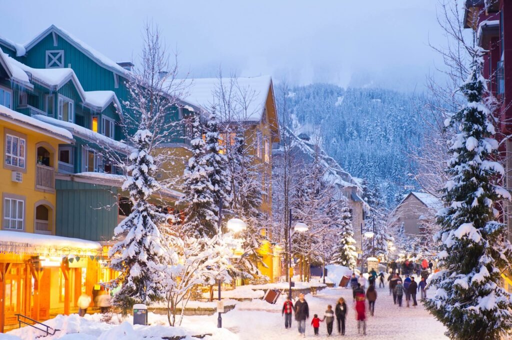 Whistler in Winter: Charming snow-covered Whistler Village at dusk with colorful buildings, glowing street lights, and people walking along the pedestrian street with mountains in background