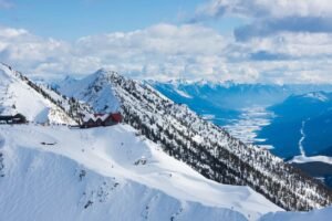 Aerial view of Kicking Horse Mountain Resort showing red-roofed alpine lodges on snowy slopes with tree-lined ski runs and Columbia Valley visible below under dramatic cloudy skies