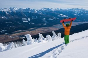 Kootenay ski resorts: Snowboarder in orange jacket and green pants holding pink snowboard overhead at mountain summit with spectacular views of Columbia River Valley, snow-covered ghost trees, and Selkirk mountain ranges at Revelstoke Mountain Resort