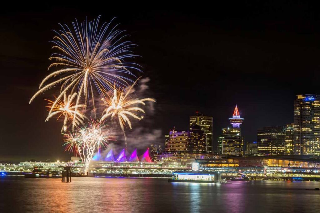 Colorful fireworks exploding over Vancouver's illuminated waterfront on New Year's Eve, with Canada Place's iconic white sails and city skyline reflecting in the harbor waters.