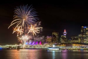 Colorful fireworks exploding over Vancouver's illuminated waterfront on New Year's Eve, with Canada Place's iconic white sails and city skyline reflecting in the harbor waters.