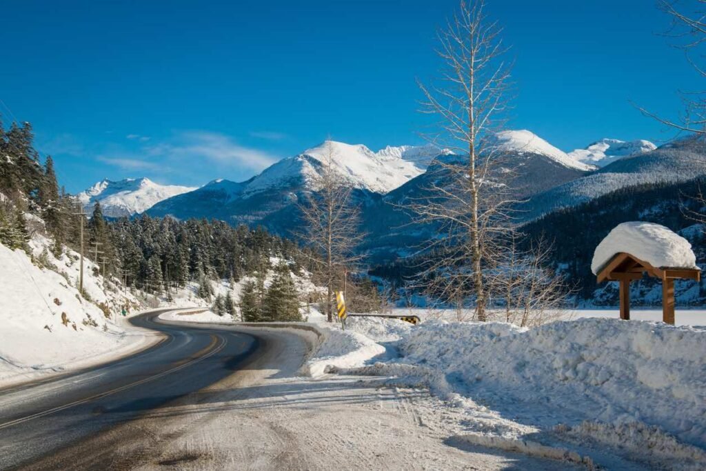 Winding highway with snow banks curving through winter landscape with snow-covered mountains and evergreen forest under blue sky along British Columbia's Sea-to-Sky Corridor