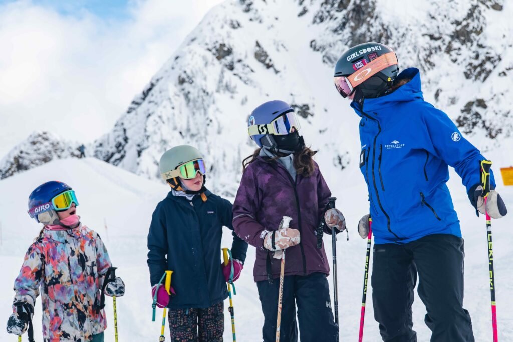 Revelstoke and Rogers Pass: A group of four people in colorful ski jackets, helmets, and goggles stand together on a snowy mountain slope, engaged in conversation. Steep, snow-covered rocky cliffs rise sharply behind them, capturing the rugged alpine atmosphere of Rogers Pass.