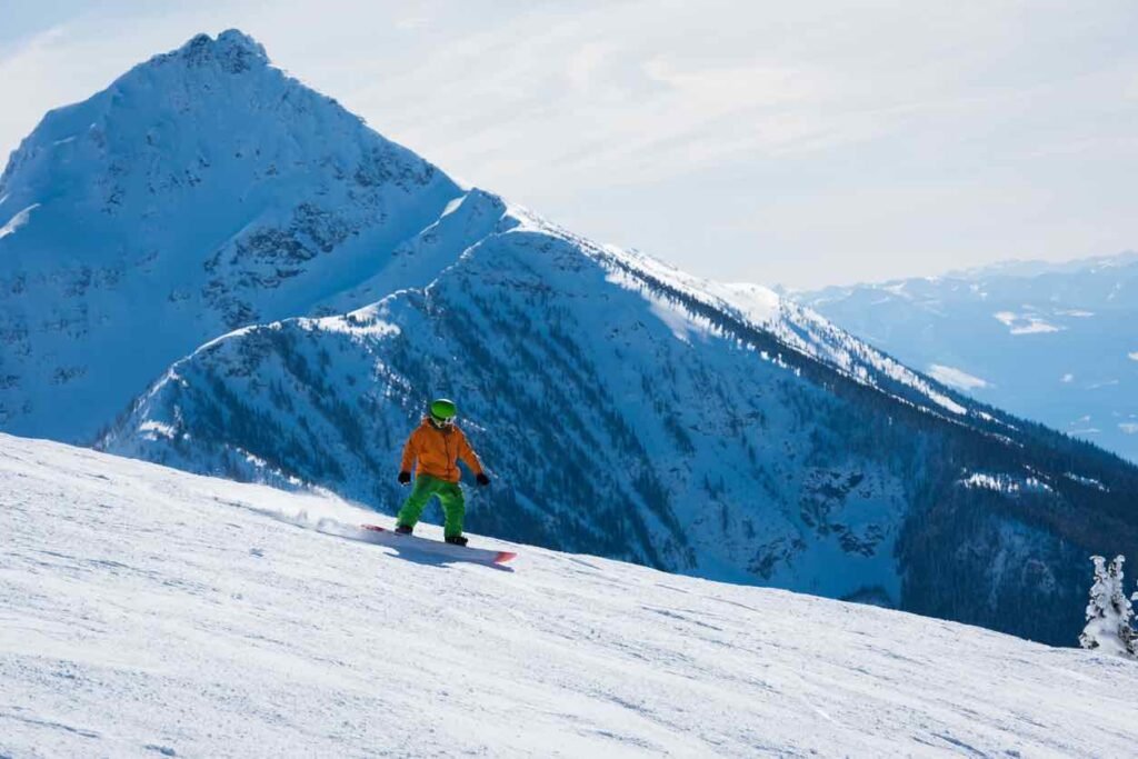 A person in a bright orange jacket and green pants snowboards down a wide, sun-lit groomed run. In the background, a massive, jagged snow-covered mountain peak rises against a clear blue sky, illustrating the vast terrain of the Selkirk Mountains.