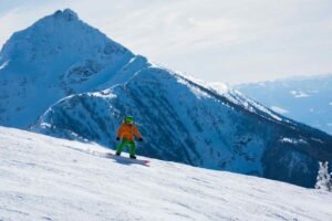 A person in a bright orange jacket and green pants snowboards down a wide, sun-lit groomed run. In the background, a massive, jagged snow-covered mountain peak rises against a clear blue sky, illustrating the vast terrain of the Selkirk Mountains.